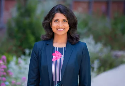 Professional outdoor portrait of a woman smiling at the camera. She is wearing a dark blazer over a striped top with a pink floral design. The background is softly blurred, featuring greenery, flowers, and a building, creating a natural campus setting.