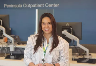 Amy Acuna stands smiling in the Peninsula Outpatient Center reception area. She is wearing a white striped blouse and staff badge, with registration desks and computer workstations visible in the background. The image highlights her role in supporting patient care and operations at the outpatient center.