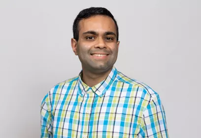 Headshot of a smiling man with short dark hair wearing a blue, green, and white plaid button-down shirt, photographed against a light gray background.