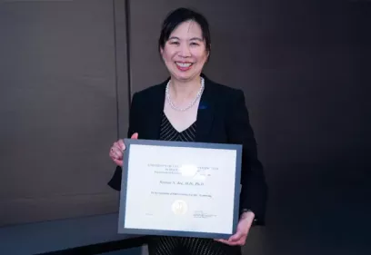 A woman wearing a dark blazer and patterned dress smiles while holding a framed certificate recognizing her outstanding faculty mentoring contributions. She stands indoors at an awards ceremony, wearing a pearl necklace, with the certificate prominently displayed.