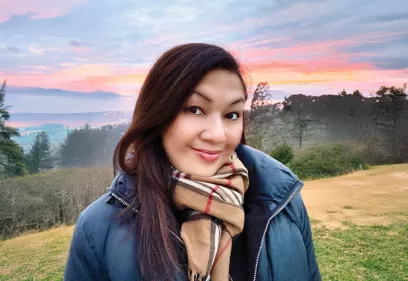 Portrait of a smiling person wearing a dark jacket and a tan plaid scarf, standing outdoors on a grassy hillside at sunrise. Trees, rolling hills, and a colorful sky create a scenic backdrop.