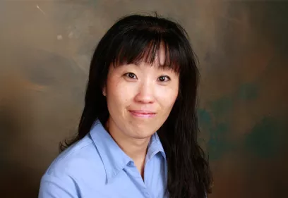 Professional portrait of a woman with long dark hair and bangs, wearing a light blue collared shirt. She is looking at the camera with a slight smile. The background is a softly blended studio backdrop in shades of brown and green, creating a warm and professional appearance.