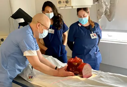 Three masked healthcare professionals in a clinical training room observe a hands-on demonstration using an anatomical head model positioned on an examination table. One instructor adjusts the model while two staff members watch. Medical imaging equipment and clinical supplies are visible in the background.