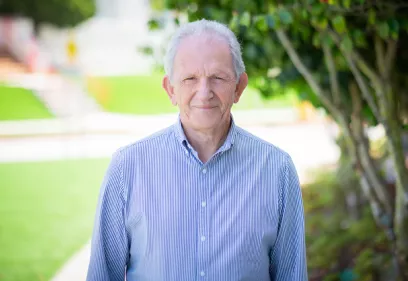 Portrait of David Saloner standing outdoors on the UCSF campus. He is wearing a blue-and-white striped button-down shirt and smiling at the camera. Trees and a sunlit walkway are softly blurred in the background. Saloner is a longtime UCSF researcher whose work has advanced vascular and cardiac imaging, including studies of atherosclerosis, aneurysms, stroke, and pulsatile tinnitus.