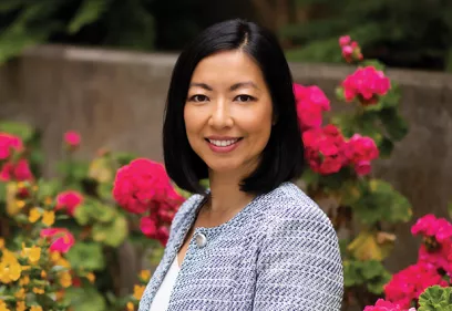 Portrait of a woman with shoulder-length dark hair smiling at the camera outdoors. She wears a patterned jacket and necklace, with vibrant pink flowers and greenery softly blurred in the background.