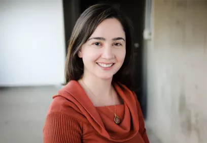 Professional portrait of Duygu Tosun-Turgut smiling at the camera. She is wearing a rust-colored sweater and a pendant necklace. The photo is taken in an outdoor or architectural setting with a softly blurred background, creating a warm and approachable appearance.