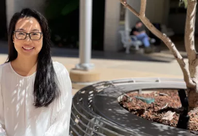 Portrait of a smiling woman with long dark hair and glasses seated outdoors in a sunlit courtyard. She is wearing a white blouse and looking toward the camera. Dappled shadows from nearby trees fall across her face and clothing. In the foreground is a circular bench surrounding a tree, while the background is softly blurred with building columns, walkways, and a person seated in the distance.