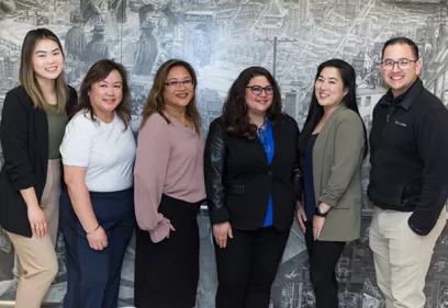 Group portrait of six smiling professionals standing side by side in front of a large black-and-white mural. The group includes five women and one man dressed in business or business-casual attire, with blazers, blouses, and collared shirts. They are facing the camera and posing for a professional photo, with the mural providing a detailed urban backdrop.