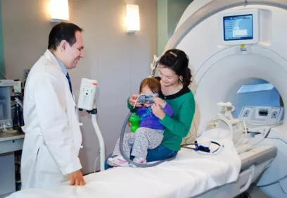 A physician stands beside a young child and her caregiver in an MRI suite. The caregiver sits on the MRI table holding the child, who is wearing special goggles and holding a green cup. The MRI scanner is visible in the background, and the interaction appears calm and reassuring as the medical team helps prepare the child for imaging.