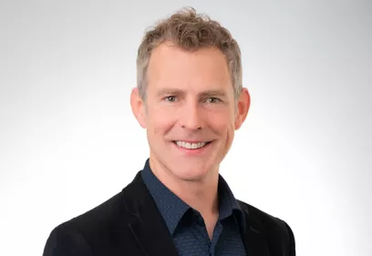 Professional headshot of a smiling man with short, curly light-brown hair wearing a dark blazer over a blue collared shirt. He is facing the camera against a plain light gray background.