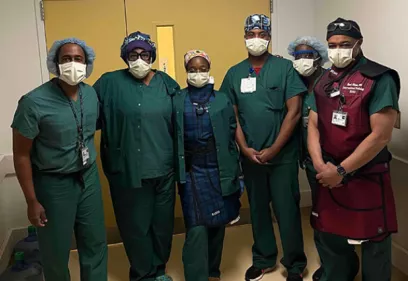 Six healthcare professionals wearing surgical scrubs, masks, and protective caps stand together in a hospital hallway outside an operating room. One team member is wearing a lead apron, and the group is facing the camera for a team photo.