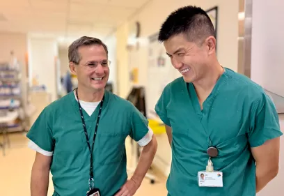 Jason Talbott and Allen Ye, both wearing green surgical scrubs, smile and talk while standing in a hospital hallway. The candid moment captures collegiality and teamwork in a clinical care setting.