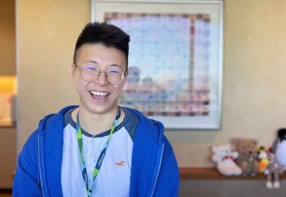 Smiling person wearing round glasses, a blue zip-up hoodie, and a conference lanyard, standing indoors in front of framed artwork. Plush toys are displayed on a shelf in the softly blurred background.
