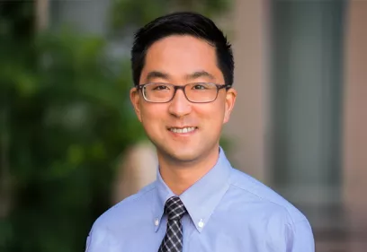 Professional headshot of a smiling individual wearing glasses, a light blue dress shirt, and a patterned tie. The portrait is framed from the chest up, with the subject facing the camera against a softly blurred outdoor background featuring greenery and architectural elements. The image conveys a friendly, approachable, and professional appearance suitable for an academic, medical, or institutional profile.