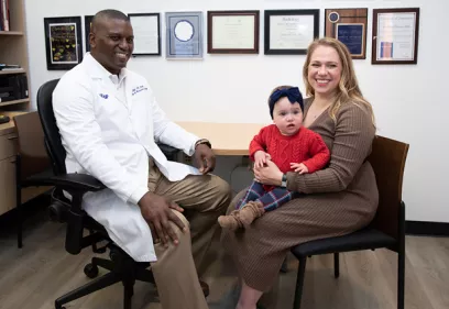 UCSF radiologist Kevin McGill sits with patient Maryana Kessel and her young child in a clinic office. McGill is wearing a white medical coat, while Kessel holds her child on her lap. Framed certificates and awards are displayed on the wall behind them, highlighting a patient-centered care setting.