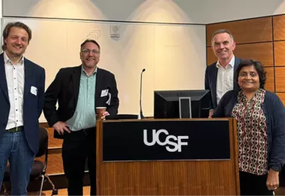 Four conference participants stand together beside a UCSF podium in a lecture hall or conference room. The group is smiling toward the camera, with the UCSF logo prominently displayed on the podium. Presentation equipment, including a microphone and monitor, is visible in the background, highlighting a professional academic or research event.