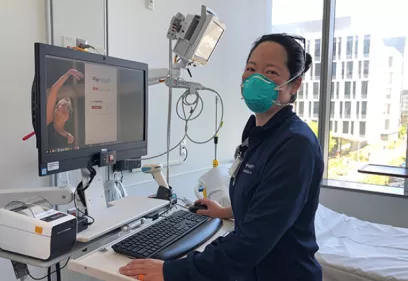A healthcare worker stands at a bedside computer workstation in a hospital patient room, smiling toward the camera while wearing a protective face mask. Medical equipment, a hospital bed, and large windows with a city view are visible in the background. A computer monitor displays a patient-related interface.