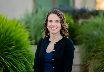 Portrait of Kimberly Kallianos, MD, smiling outdoors, seated in front of softly blurred green landscaping.
