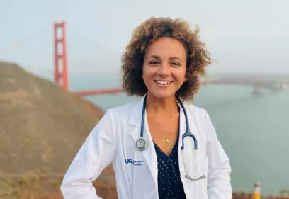 Portrait of a smiling person with curly hair wearing a white UCSF School of Medicine lab coat and a stethoscope over a dark patterned top. They are standing outdoors with the Golden Gate Bridge and San Francisco Bay softly blurred in the background.