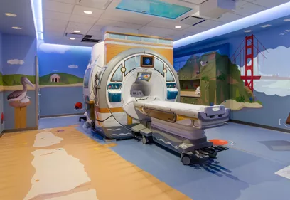 A brightly decorated pediatric MRI suite with a colorful beach and San Francisco-themed mural surrounding the scanner. The MRI machine is designed with child-friendly artwork, and the room features playful illustrations to create a welcoming environment for young patients.