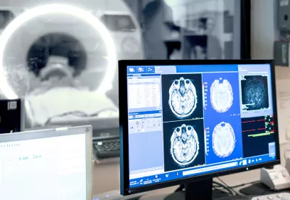 A radiology workstation displays brain MRI scans and imaging data as a patient is positioned inside an MRI scanner visible through a control room window.
