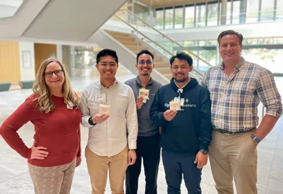 Five individuals stand together in a bright, modern lobby holding small cards or badges and smiling toward the camera. The group is casually dressed, with one person wearing a UCSF Benioff Children's Hospitals sweatshirt. A staircase and large windows are visible in the background, creating an open and welcoming environment. The photo appears to have been taken during a workplace event, training activity, or recognition program.