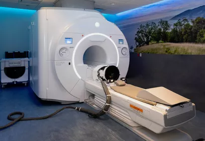 Wide view of an MRI scanner with the patient table extended in a modern imaging suite. The room features blue accent lighting and a large wall mural of a grassy landscape with trees and mountains, creating a calming environment.
