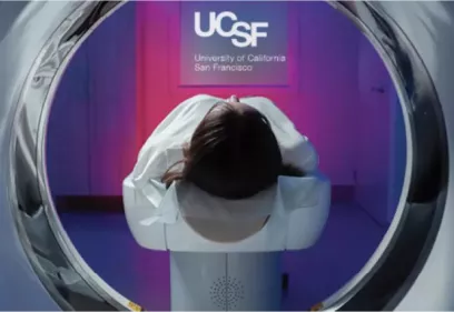 A patient lies on an MRI table positioned inside the opening of an MRI scanner at UCSF. The image is viewed from inside the scanner bore, with purple lighting and UCSF branding visible in the background. The scene illustrates a patient undergoing an MRI examination in a modern imaging facility.