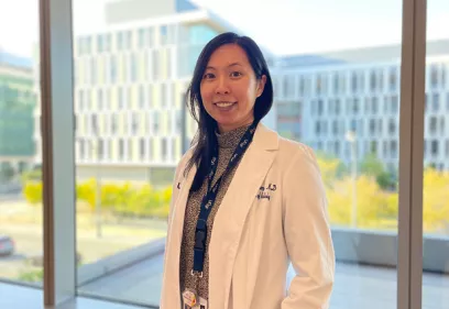 Professional portrait of a physician standing indoors in front of large windows overlooking a modern campus. She is wearing a white lab coat, an identification badge, and a dark patterned top, and is smiling at the camera. The background features softly blurred buildings and trees in daylight.