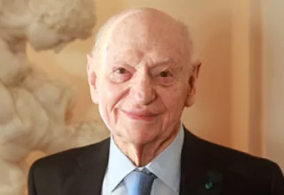 Portrait of an elderly man wearing a dark suit jacket, light blue dress shirt, and blue tie. He is smiling gently at the camera in a warmly lit indoor setting. A classical marble sculpture is visible in the softly blurred background, adding an elegant and distinguished atmosphere to the portrait.