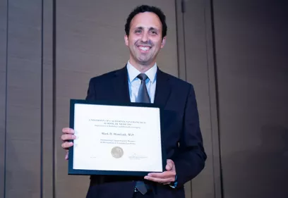 Mark Mamlouk stands in a suit and tie, smiling while holding a framed UCSF certificate recognizing his service as a volunteer physician. He is photographed indoors against a neutral background during an awards ceremony.