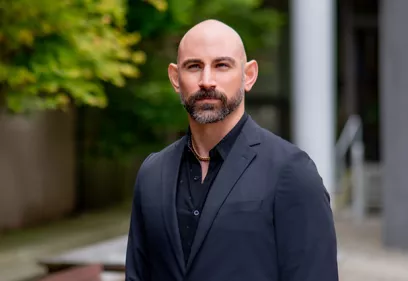 Professional portrait of a man with a shaved head and neatly trimmed beard wearing a dark suit jacket over a black shirt. He stands outdoors with a serious expression, while greenery and modern building elements are softly blurred in the background.