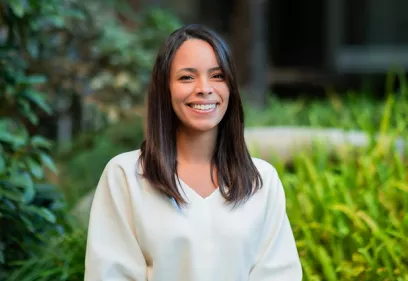 Smiling woman with shoulder-length dark hair wearing a cream-colored blouse, standing outdoors in front of softly blurred greenery and landscaping. She is facing the camera with a friendly expression in a natural garden setting.