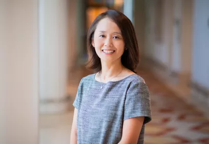 Professional portrait of a woman with shoulder-length dark hair wearing a gray short-sleeved dress and a delicate necklace. She is smiling at the camera while standing in a softly lit indoor hallway, with columns and a patterned floor blurred in the background.