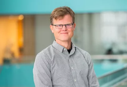 Portrait of Peder Larson wearing glasses and a checkered button-down shirt, smiling at the camera. He stands indoors with a softly blurred modern building interior in the background.