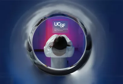 View from inside the circular opening of an MRI scanner looking toward a patient positioned on the imaging table. The patient lies face down with their head centered in the scanner. Beyond the scanner opening, a softly illuminated UCSF sign glows against a purple and pink backdrop, creating a modern, futuristic atmosphere. The image emphasizes advanced medical imaging technology and the patient experience within a radiology environment.