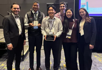 Group of six radiology trainees and professionals posing together at a conference awards event. The group stands indoors in front of a presentation stage, wearing conference badges and business or business-casual attire. The person in the center holds a large silver trophy labeled for a resident competition, while another participant holds an individual award plaque. All six are smiling toward the camera, celebrating their achievement. The image highlights teamwork, academic excellence, and recognition at a