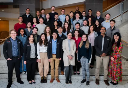 Group portrait of radiology residents, fellows, and faculty gathered on a staircase inside a medical center. The group is dressed in a mix of professional and business-casual attire and poses together in a bright atrium, highlighting the collaborative training environment within the radiology program.