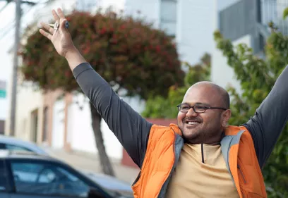 Rahul Desikan smiles and raises his arms while holding keys in one hand, standing on a city street in front of trees and residential buildings.