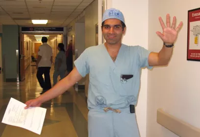 A healthcare professional wearing surgical scrubs and a surgical cap stands in a hospital hallway, smiling and waving at the camera. He holds a document in one hand while raising the other in greeting. Staff members can be seen in the background, and the clinical setting suggests a busy hospital environment.