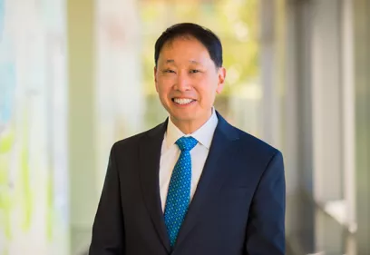 Portrait of a smiling person wearing a dark suit, white shirt, and blue patterned tie. They are standing indoors in a hallway with a softly blurred background, facing the camera.