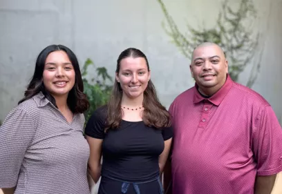 Three staff members standing outdoors and smiling at the camera in front of a concrete wall with greenery. Two women and one man are posed side by side, wearing professional casual attire. The group portrait is taken in a landscaped courtyard setting with plants and vines visible in the background.
