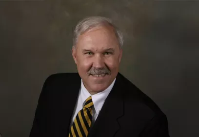 Professional headshot of a man with gray hair and a mustache, wearing a dark suit, white dress shirt, and black-and-gold striped tie. He is smiling at the camera against a softly blended studio background, creating a formal and distinguished portrait.