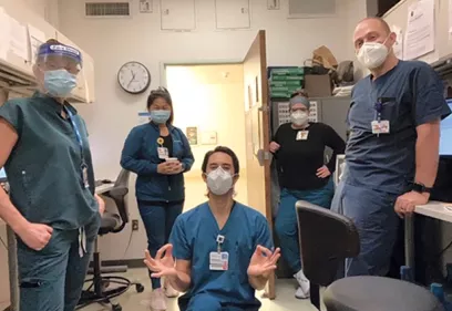 Five healthcare professionals wearing scrubs and face masks pose together in a clinical workroom. One staff member kneels in the center while the others stand around the room. Computers, office chairs, medical supplies, and a wall clock are visible in the background, highlighting a hospital or imaging department workspace. The group appears to be taking a brief team photo during their shift.