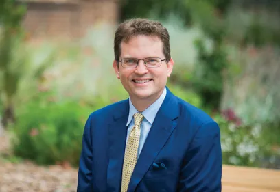 Portrait of a smiling man wearing glasses, a blue suit, and a yellow patterned tie, seated outdoors with a softly blurred garden background.