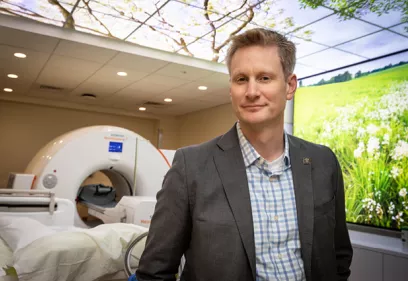 Portrait of a man standing in a modern medical imaging suite beside an MRI scanner. He is wearing a gray blazer over a blue-and-white checkered shirt and is looking directly at the camera with a slight smile. Behind him, the MRI machine and patient table are visible, while large wall and ceiling displays show bright nature scenes with green fields and tree branches. The clinical environment is designed to create a calming atmosphere for patients undergoing imaging procedures.