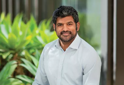 Professional outdoor portrait of a man with short dark hair and a trimmed beard, wearing a light blue button-down shirt. He is seated and smiling at the camera, with lush green plants and a modern building softly blurred in the background. Natural lighting creates a warm and approachable professional headshot.