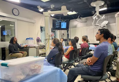 Students participate in an ultrasound-guided procedure training session in a simulation lab. An instructor demonstrates techniques using a training model while students observe imaging displayed on a monitor and ultrasound machine.