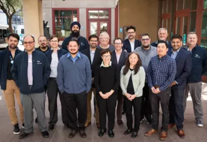 Group photo of a large radiology information technology team standing together outside a building entrance. Sixteen team members are arranged in two rows, facing the camera and smiling. They are dressed in a mix of business casual and professional attire, including sweaters, button-down shirts, jackets, and blazers. The group stands in a courtyard area in front of glass doors and large windows, with trees and urban buildings visible in the background. The image conveys teamwork, collaboration, and a profess