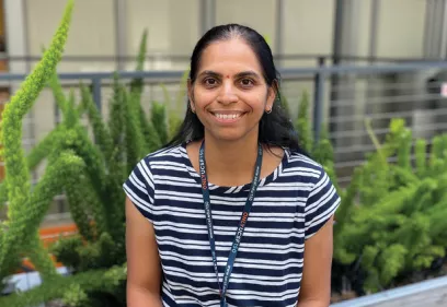 Portrait of a smiling person with long dark hair wearing a navy and white striped shirt and a UCSF lanyard. They are seated outdoors in front of green plants and a modern building with a softly blurred background.